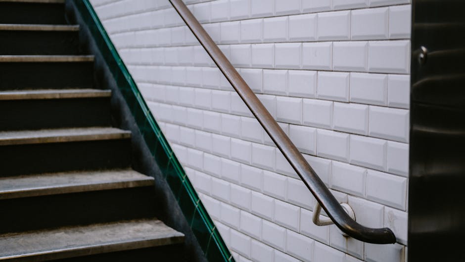 Exterior view of a concrete staircase with moss and dirt accumulation between the steps, featuring a metal handrail running along the left side. The staircase is outdoors, leading to a building with a