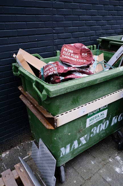 A green outdoor waste container filled with red bags of weed control products, positioned on a wheeled platform against a dark blue brick wall. The container's lid is open, revealing the bags stacked 