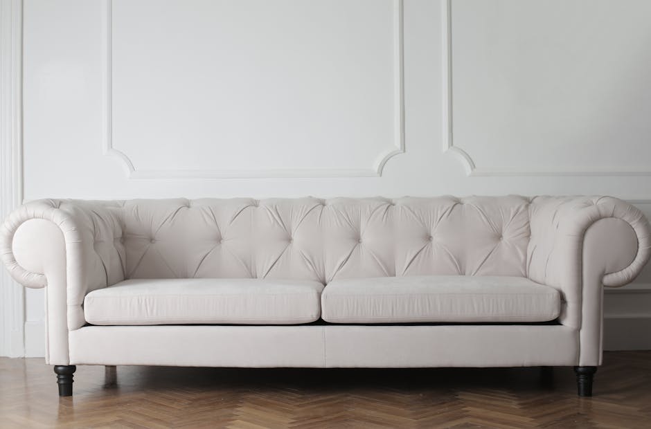 A person wearing black gloves is using a yellow handheld polymer surface cleaner with a circular cleaning pad to deep clean a dark grey fabric sofa in a residential living room. The sofa has a modern