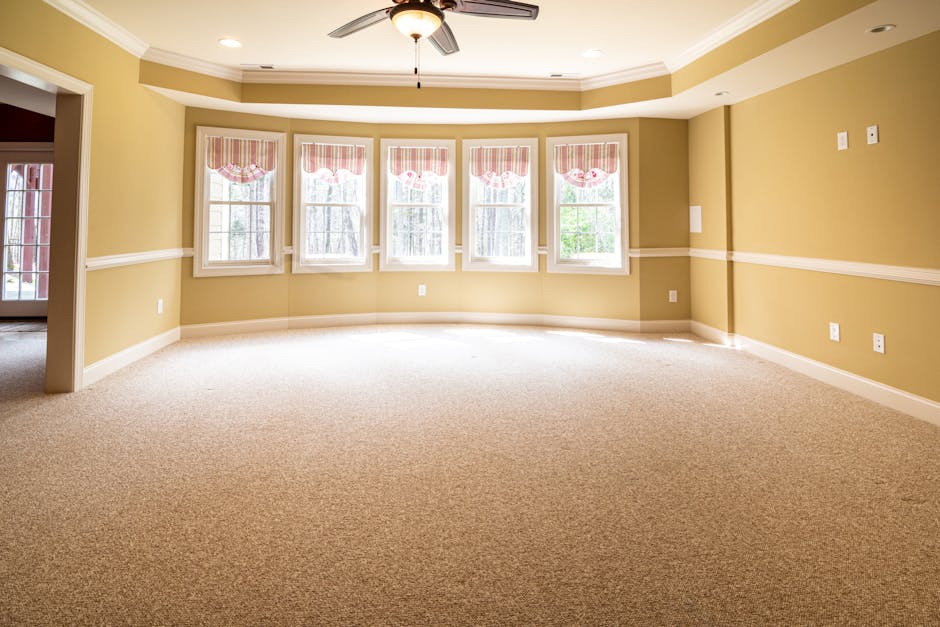 A spacious, sunlit living room with beige carpet flooring and soft yellow walls featuring white crown molding. The room has a curved wall with five large double-hung windows, each dressed with pink an