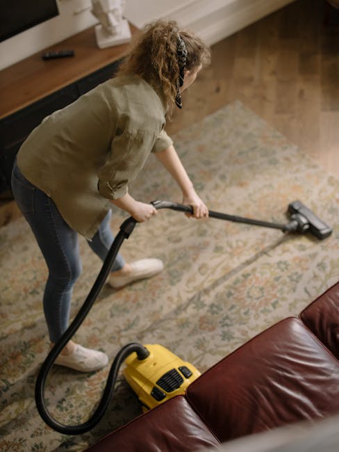 A person performing surface cleaning with a vacuum cleaner on a patterned rug inside a residential living room. The individual is wearing casual clothing, including a beige jacket, dark jeans, and whi
